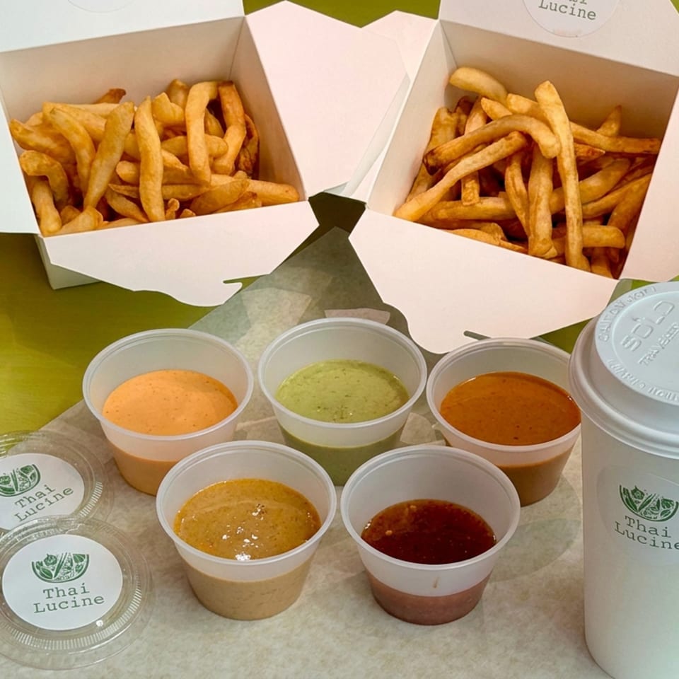 Thai Lucine takeout spread with two boxes of fries, five dipping sauces, and two iced drinks on a green table.
