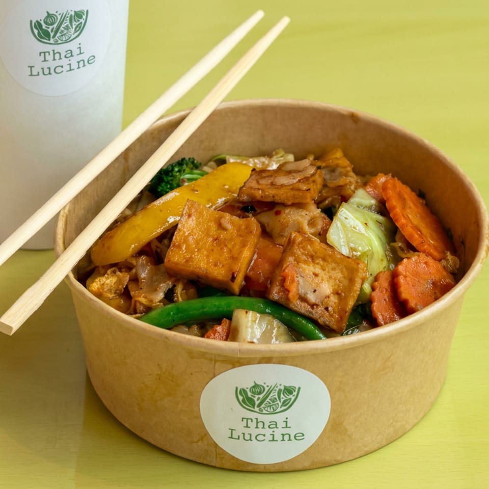 Thai Lucine takeout bowl filled with tofu, vegetables, and noodles, accompanied by chopsticks and a branded drink cup.