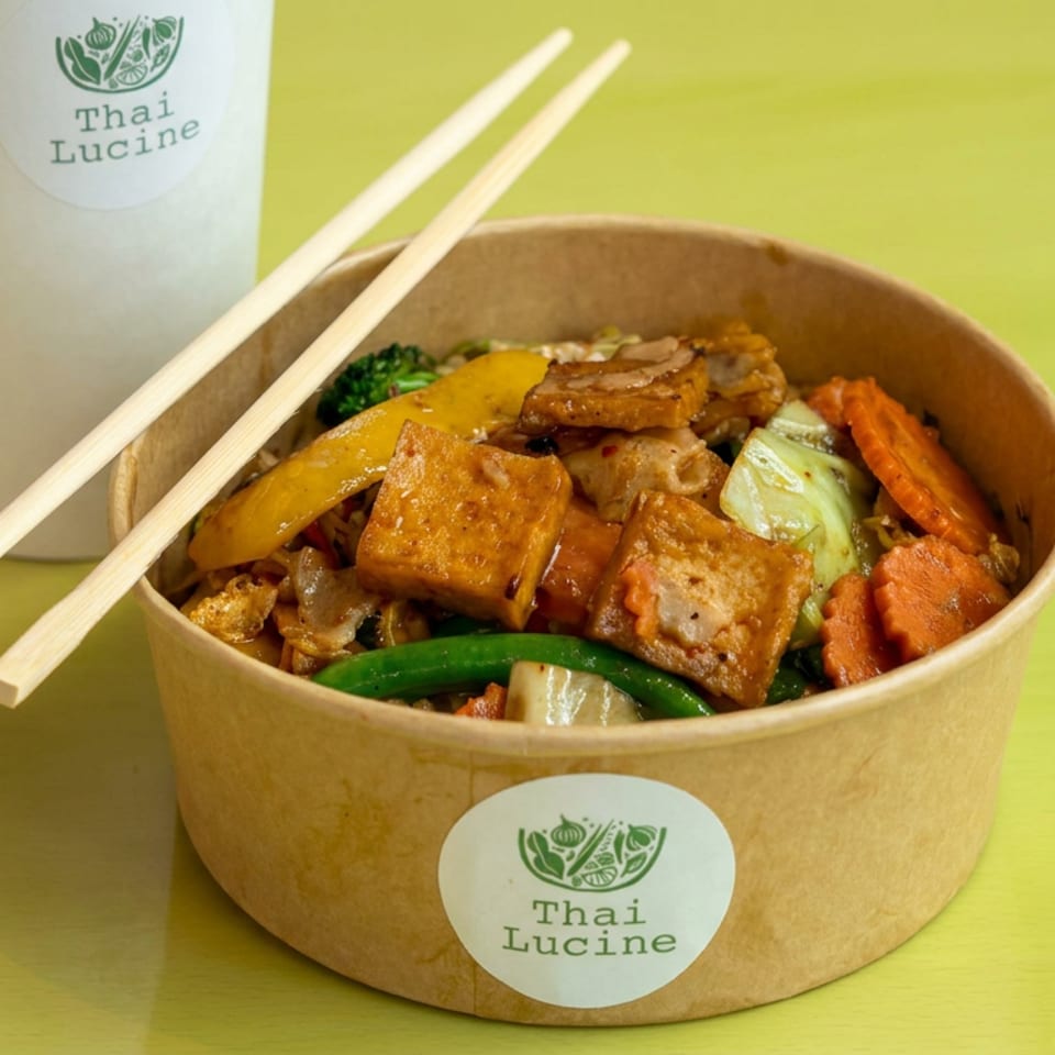 Thai Lucine bowl of stir-fried vegetables and tofu with chopsticks, accompanied by a branded drink on a bright green table.